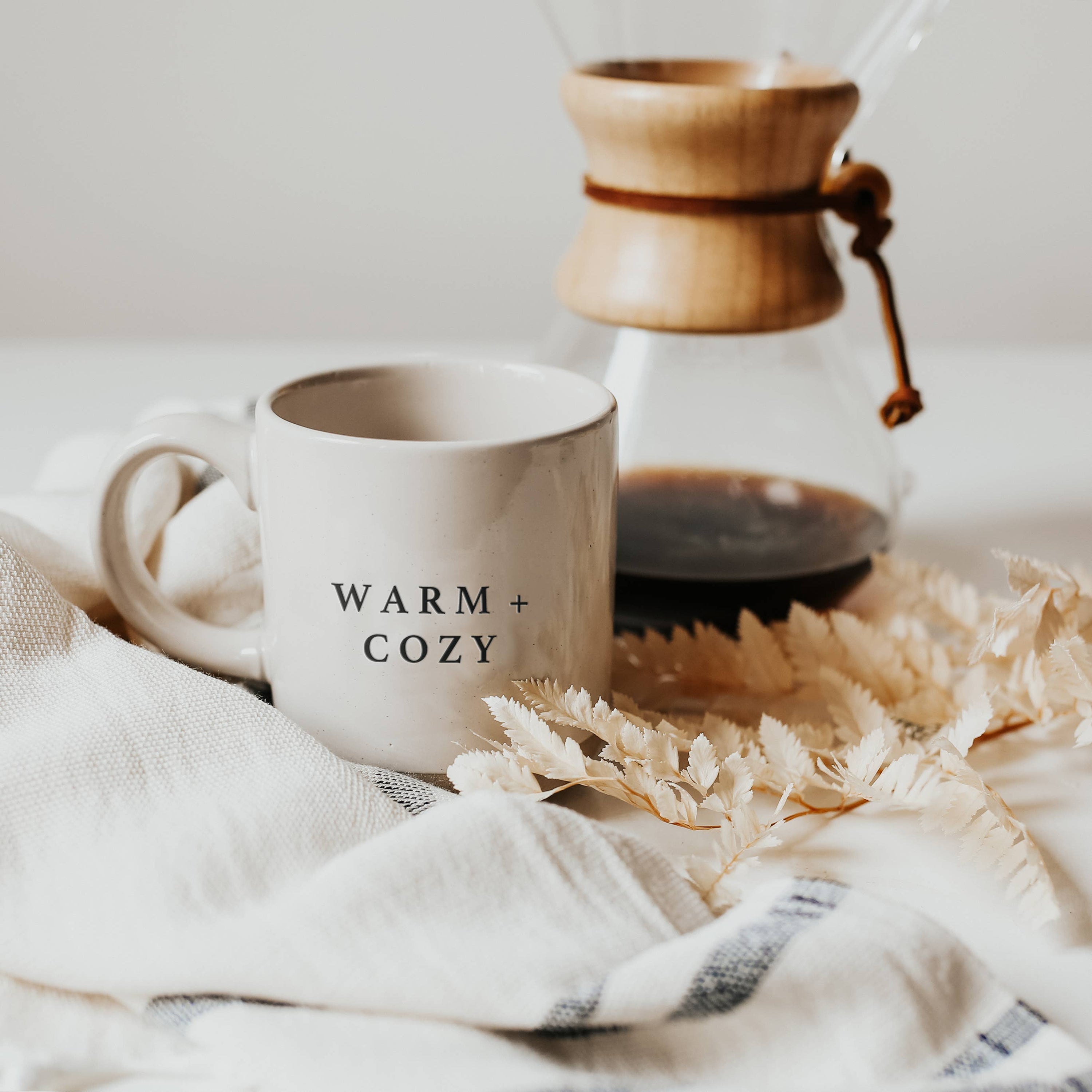 White mug with 'WARM + COZY' text next to a glass carafe on a soft surface.