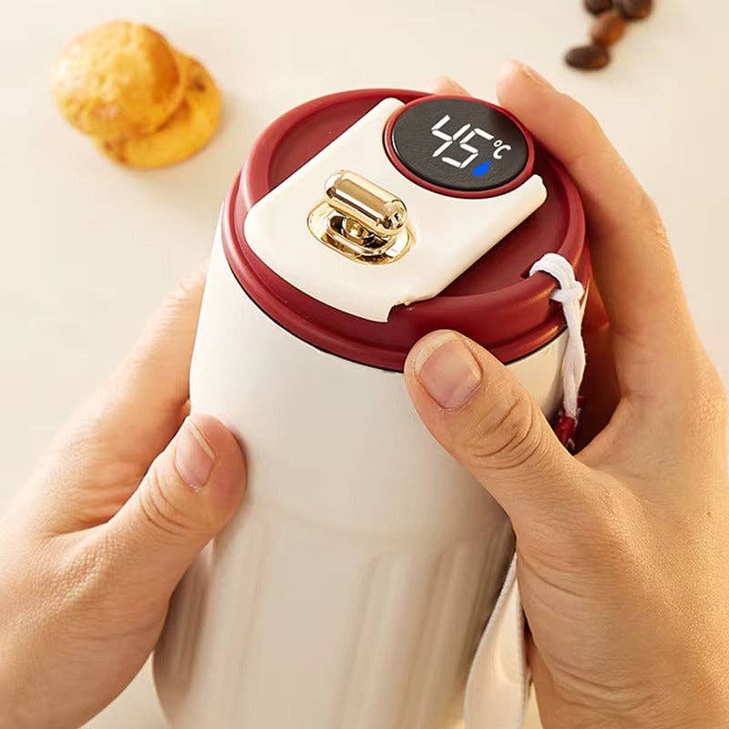 White mug with red lid and digital temperature display held by hands, with a pastry in the background.