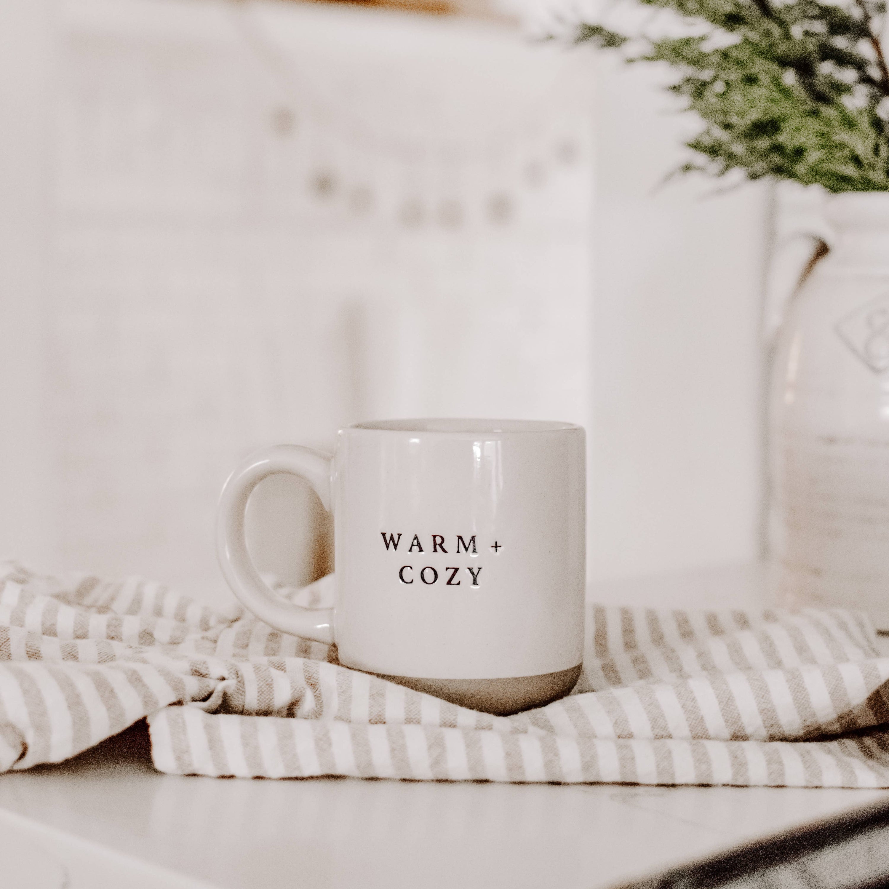 White mug with 'WARM + COZY' text on a striped blanket with a blurred background