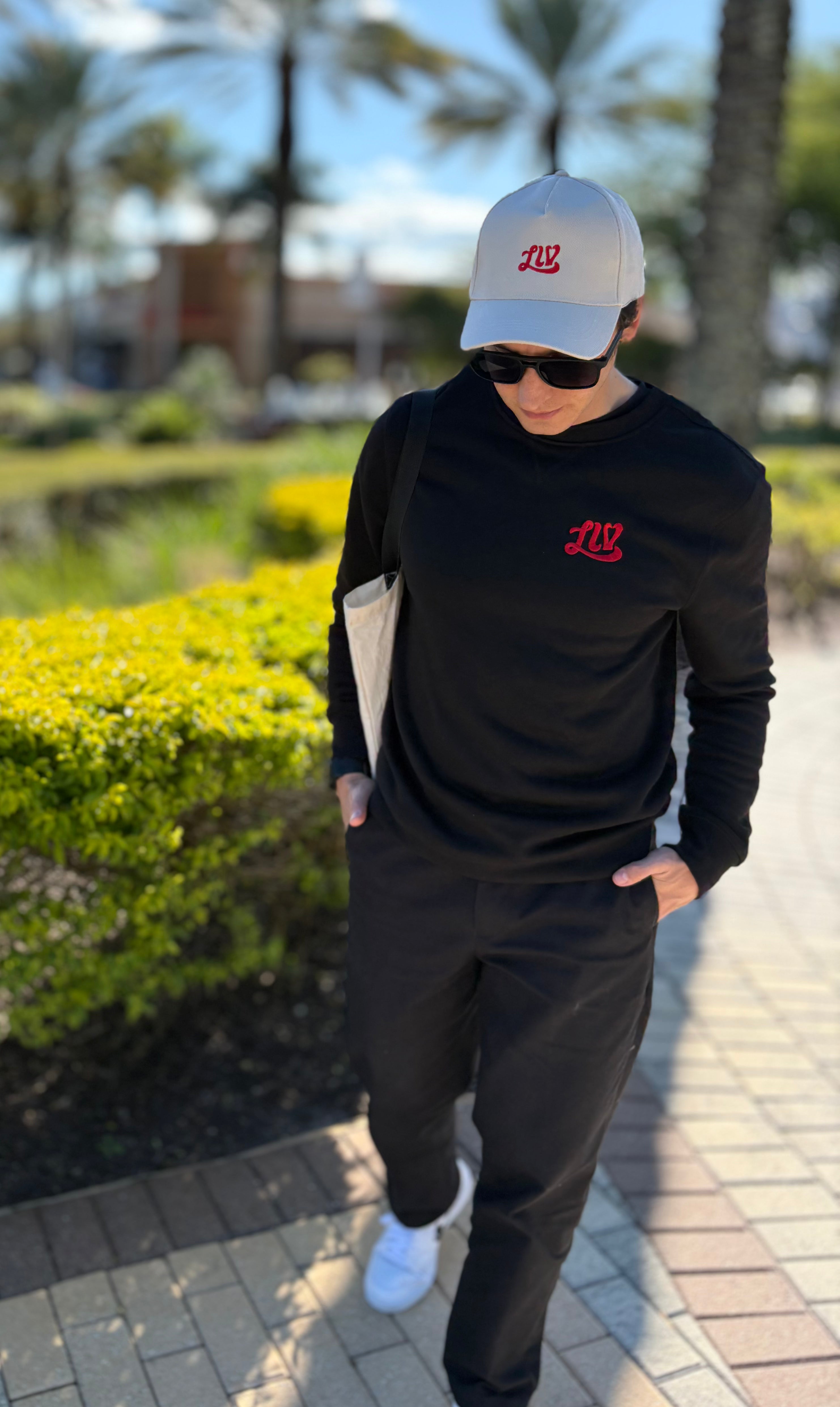Person wearing a black hoodie with a red logo, white cap, and sunglasses, walking outdoors.