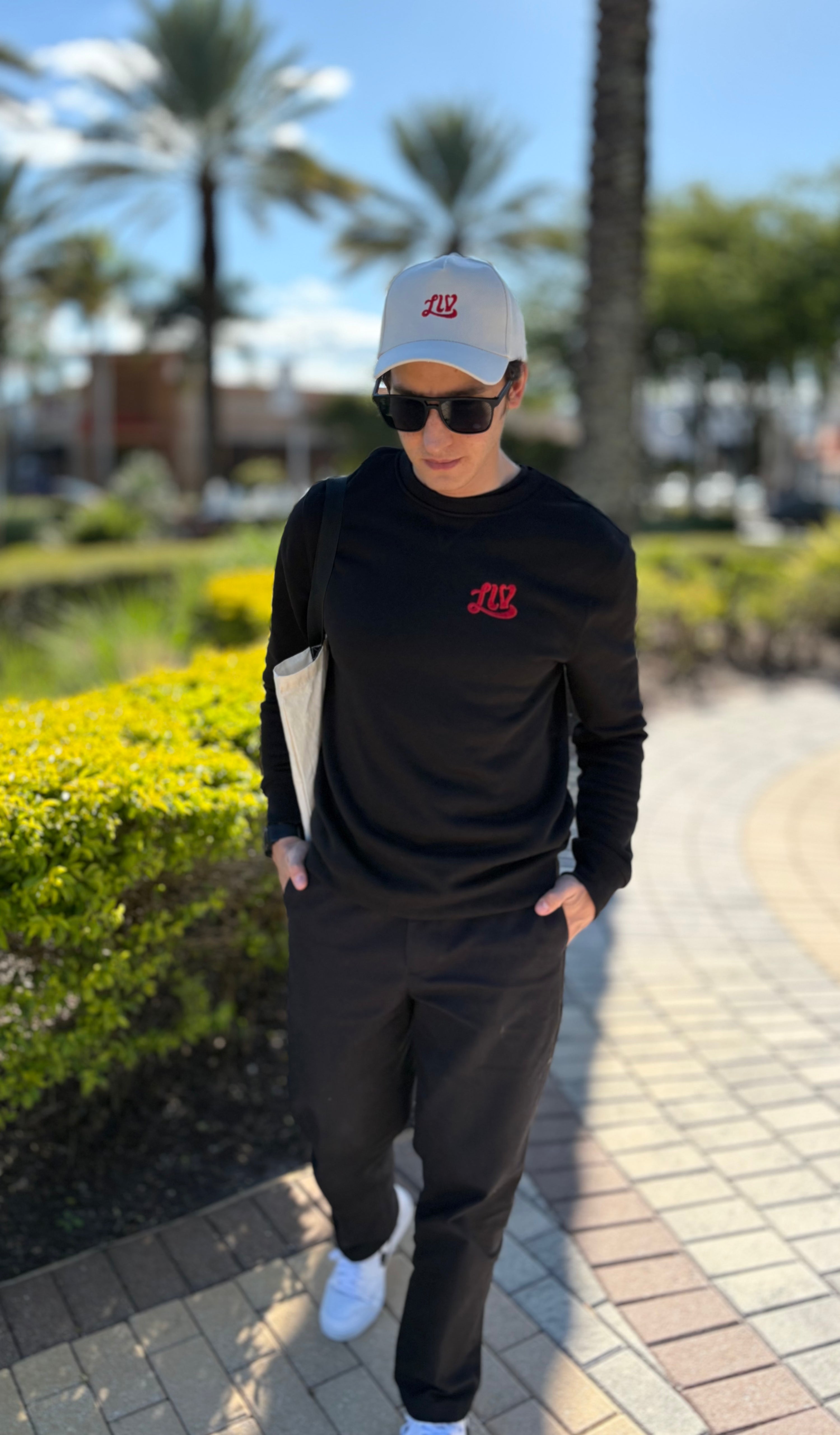 Person wearing a black outfit with logos, sunglasses, and a cap in an outdoor setting with palm trees.