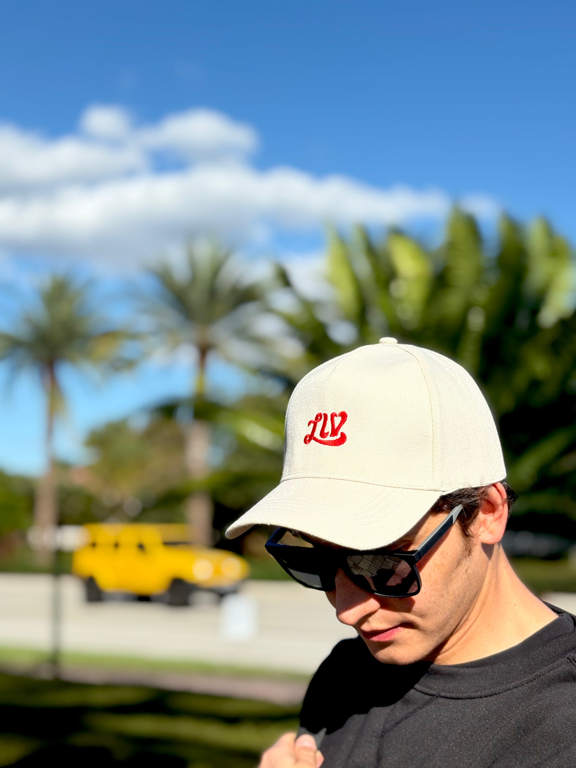 Man wearing a beige cap with a red logo and sunglasses, standing outdoors with palm trees in the background.