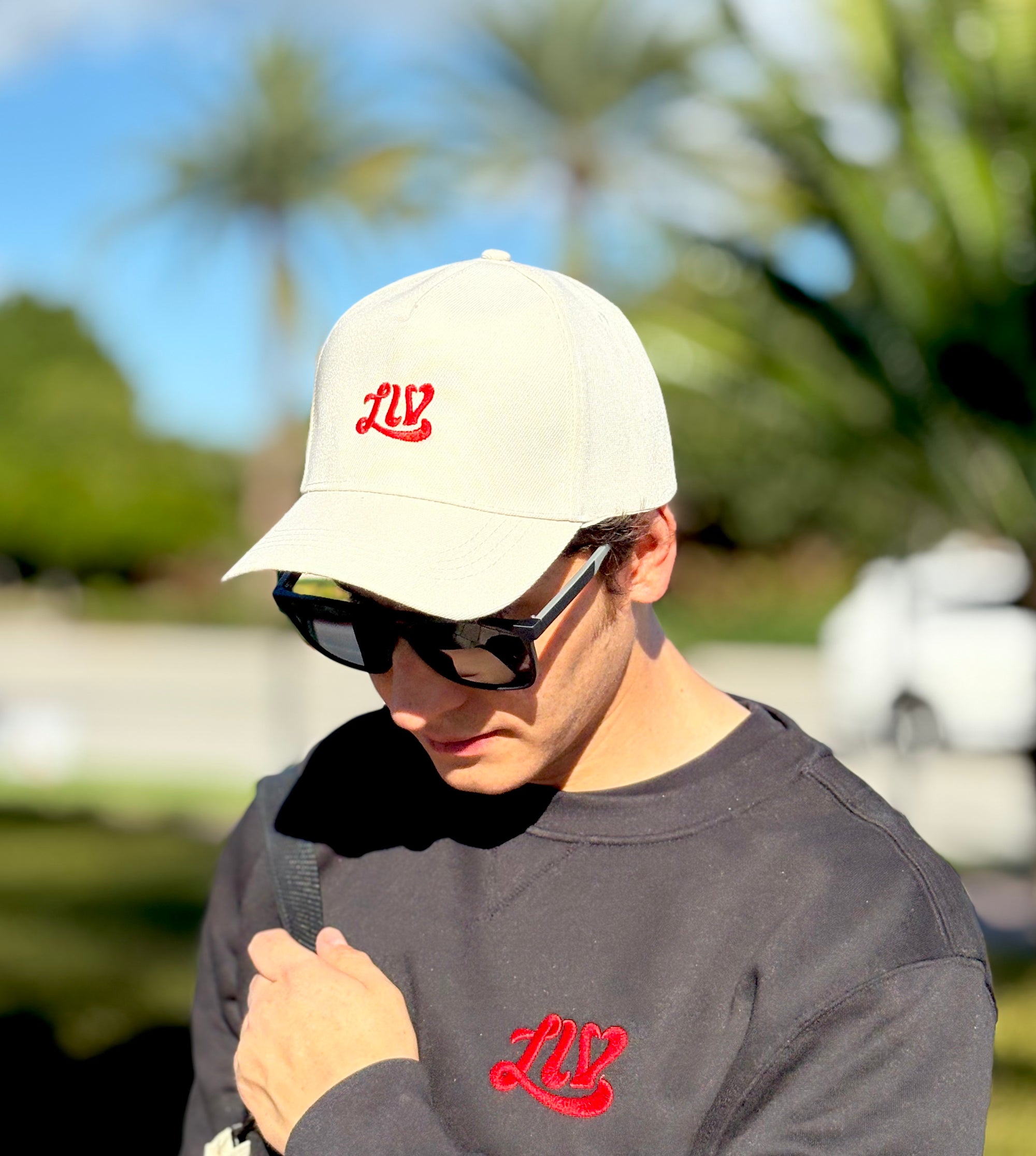 Man wearing a beige cap and sunglasses with a red logo, standing outdoors.