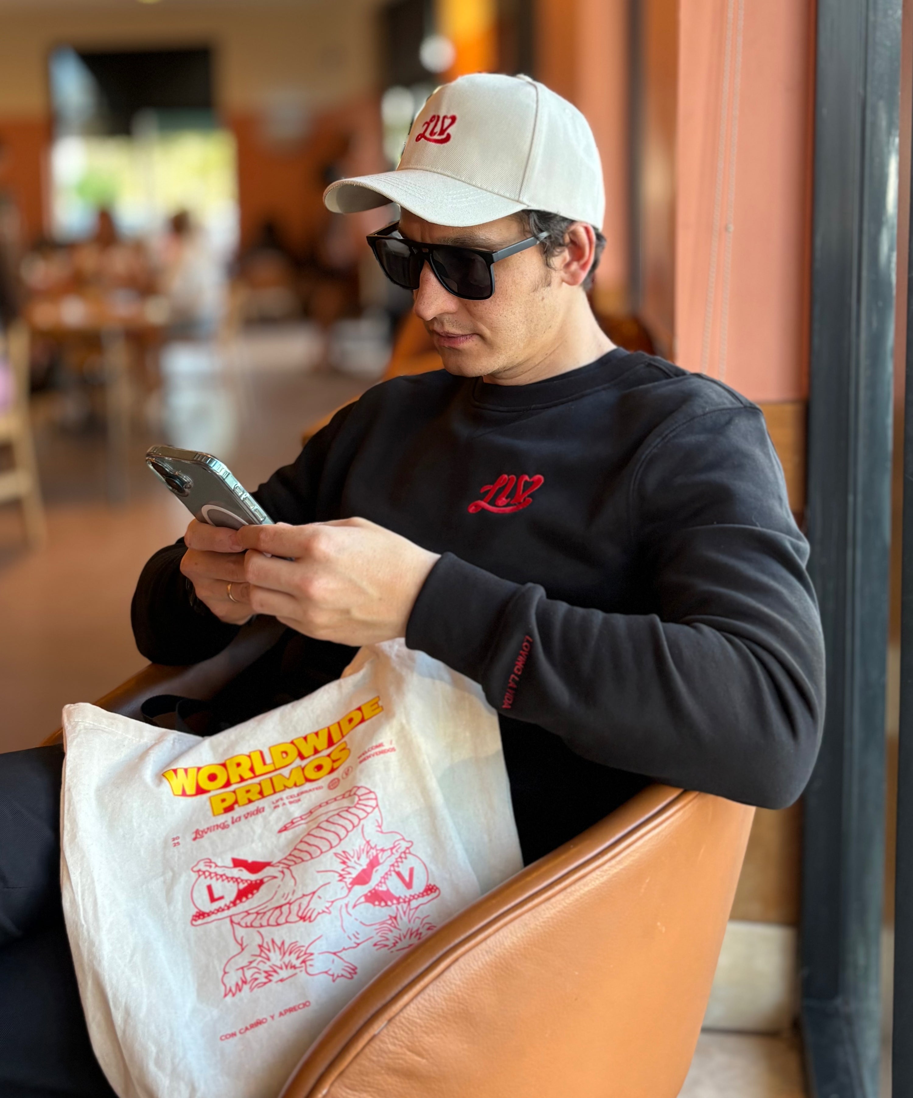 Man wearing a cap and sunglasses, using a phone, with a branded bag in a casual setting.