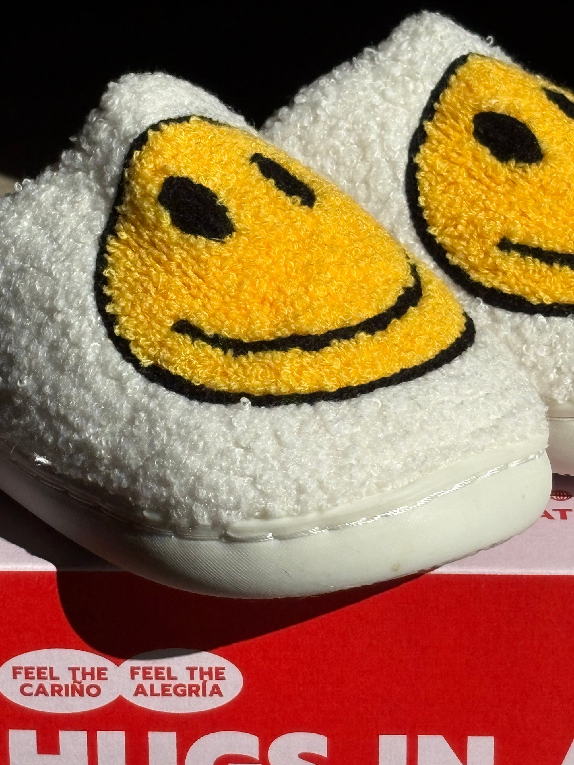 White slippers with yellow smiley face design on a red box background