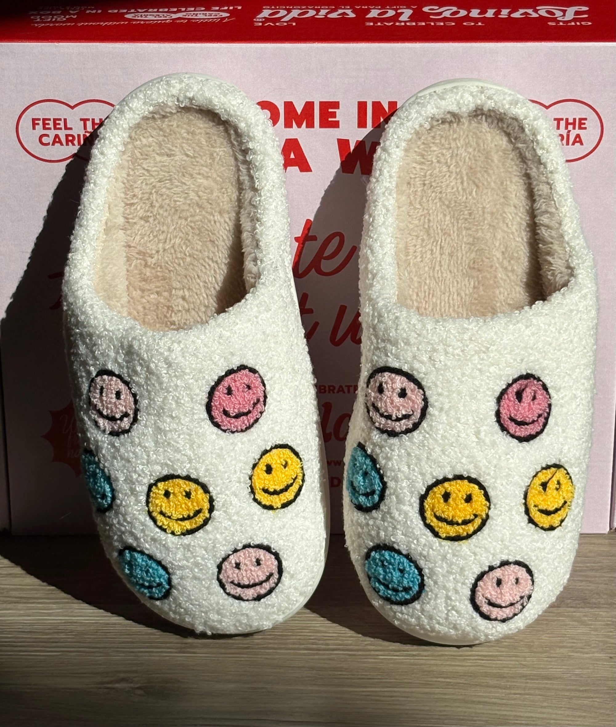 Pair of white slippers with colorful smiley face patterns on a wooden surface.