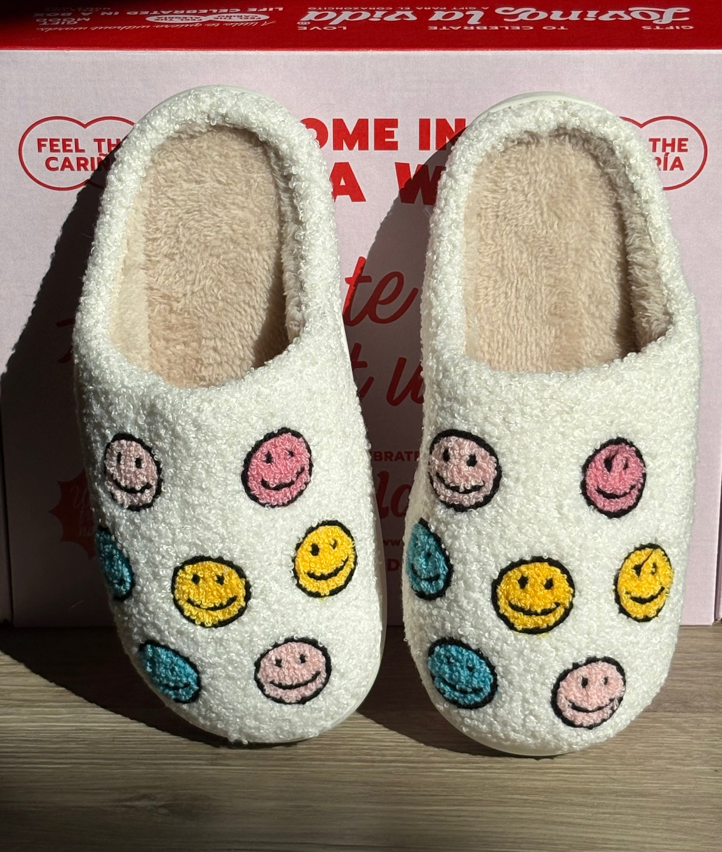 Pair of white slippers with colorful smiley face patterns on a wooden surface.