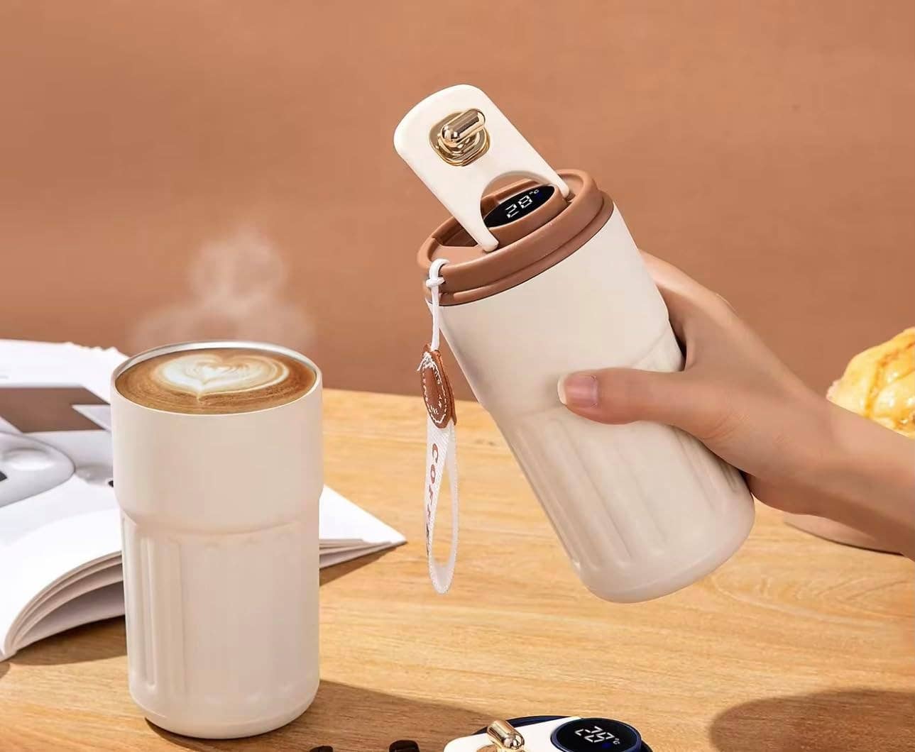 Hand holding a white travel mug with a digital display, next to a steaming cup of coffee on a wooden table.