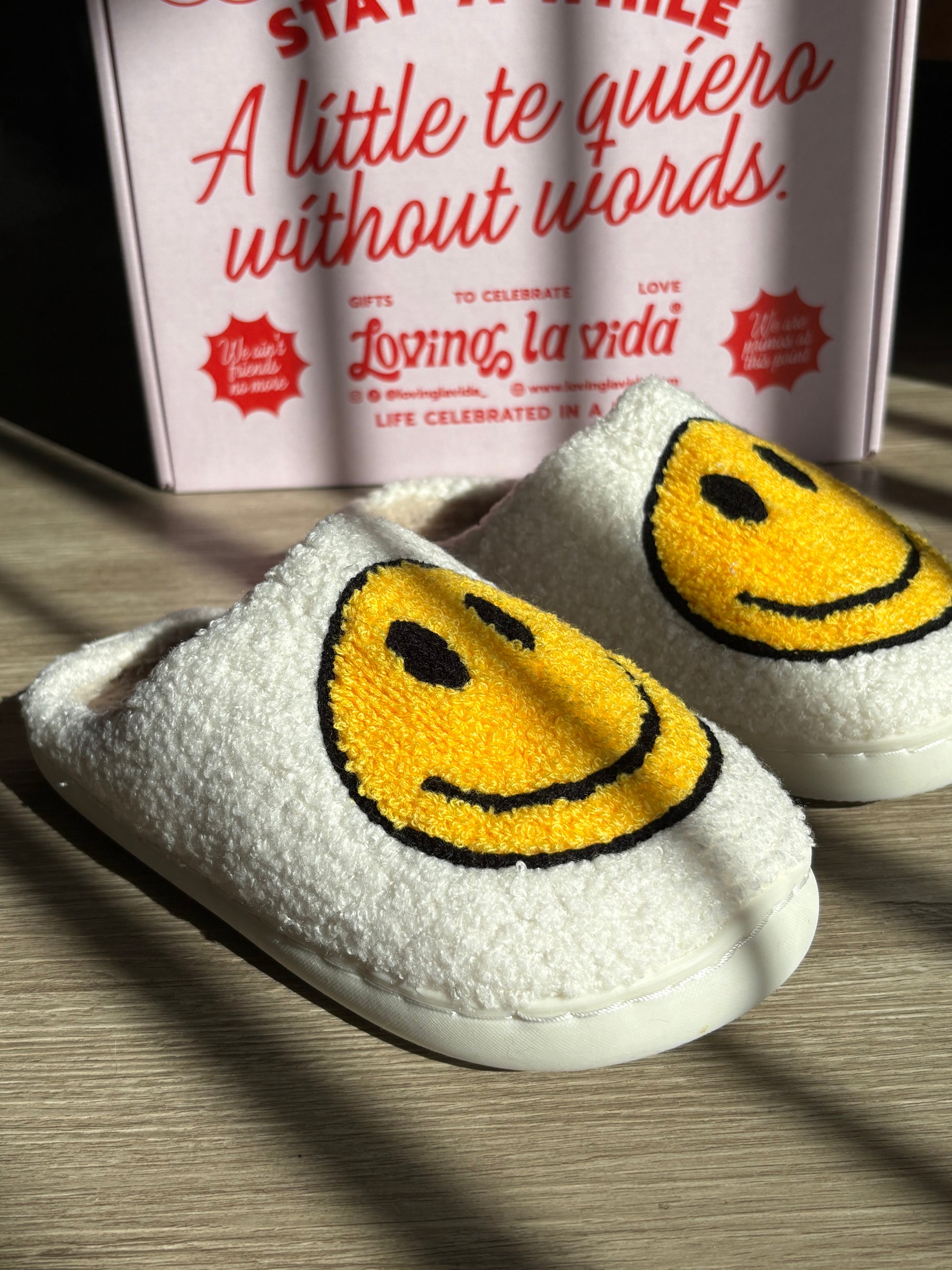 White slippers with yellow smiley faces on a wooden surface, with a product box in the background.